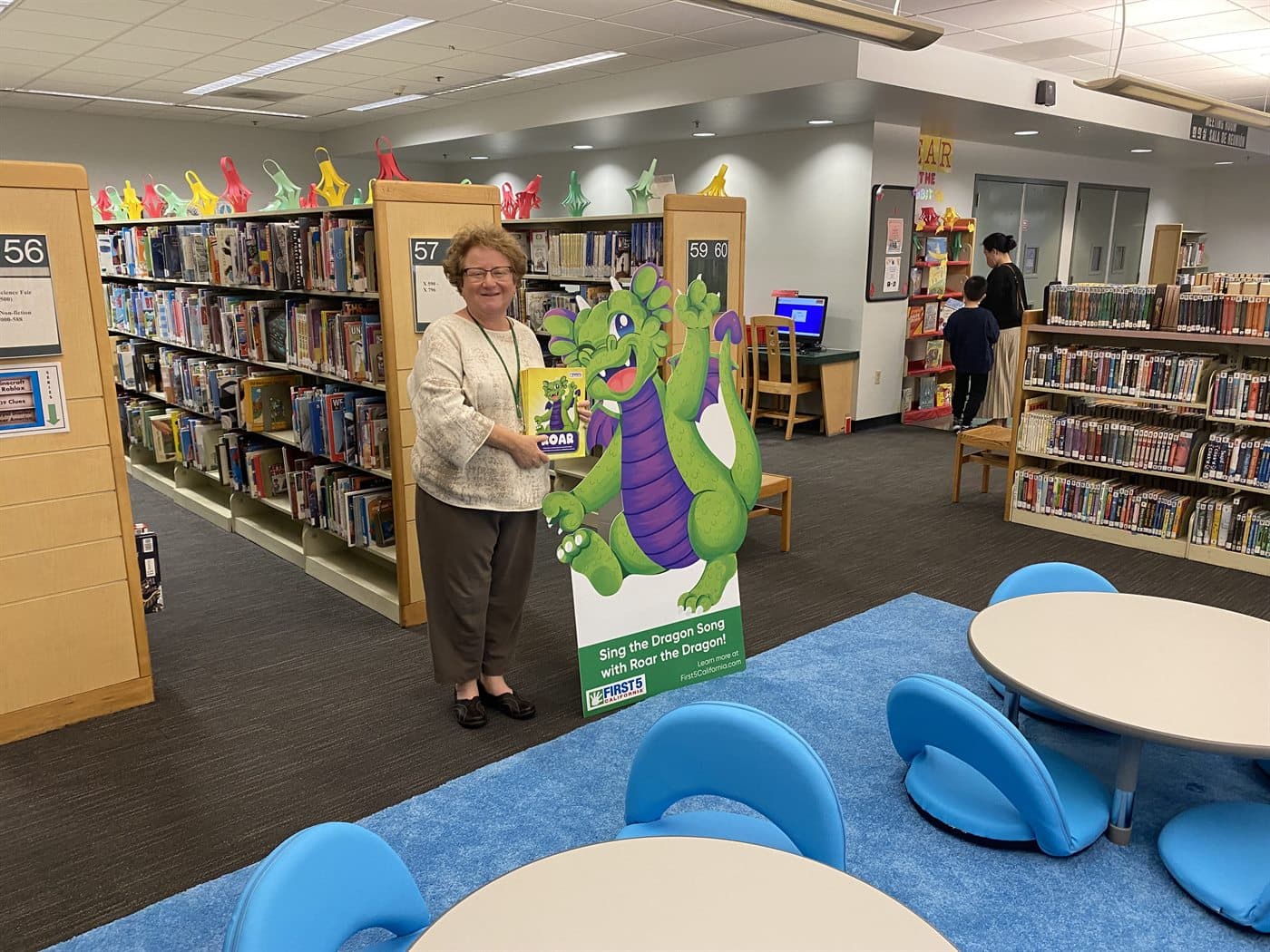 A librarian standing beside a children's reading-program standee among the shelves of a public library