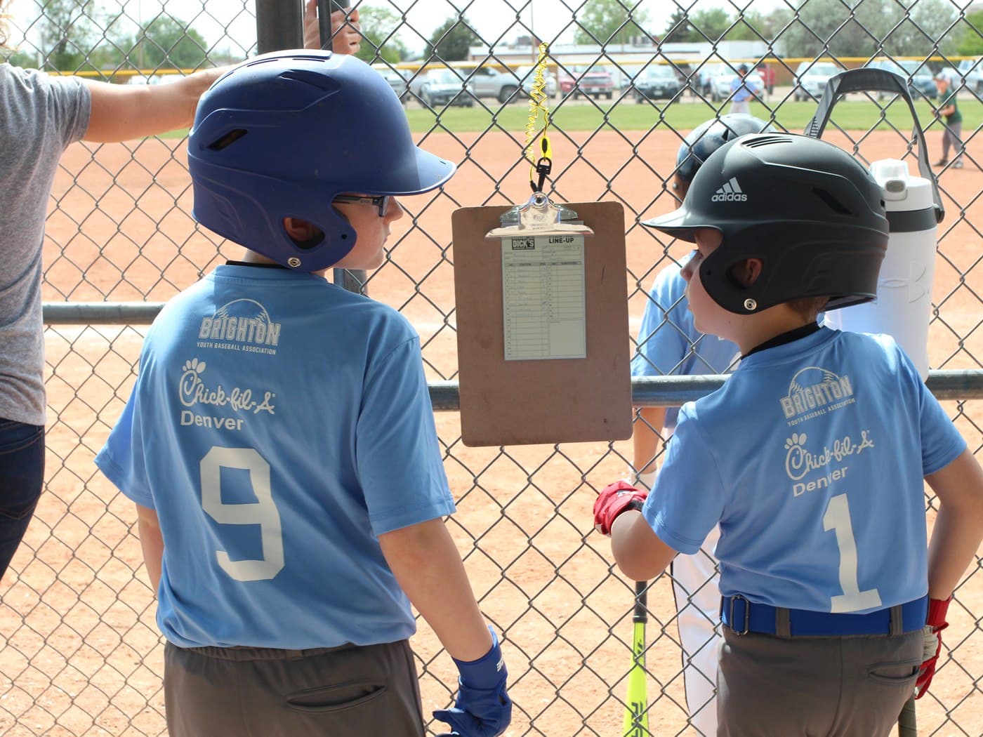 Two young baseball players in caps reading a posted community message at a ballfield fence