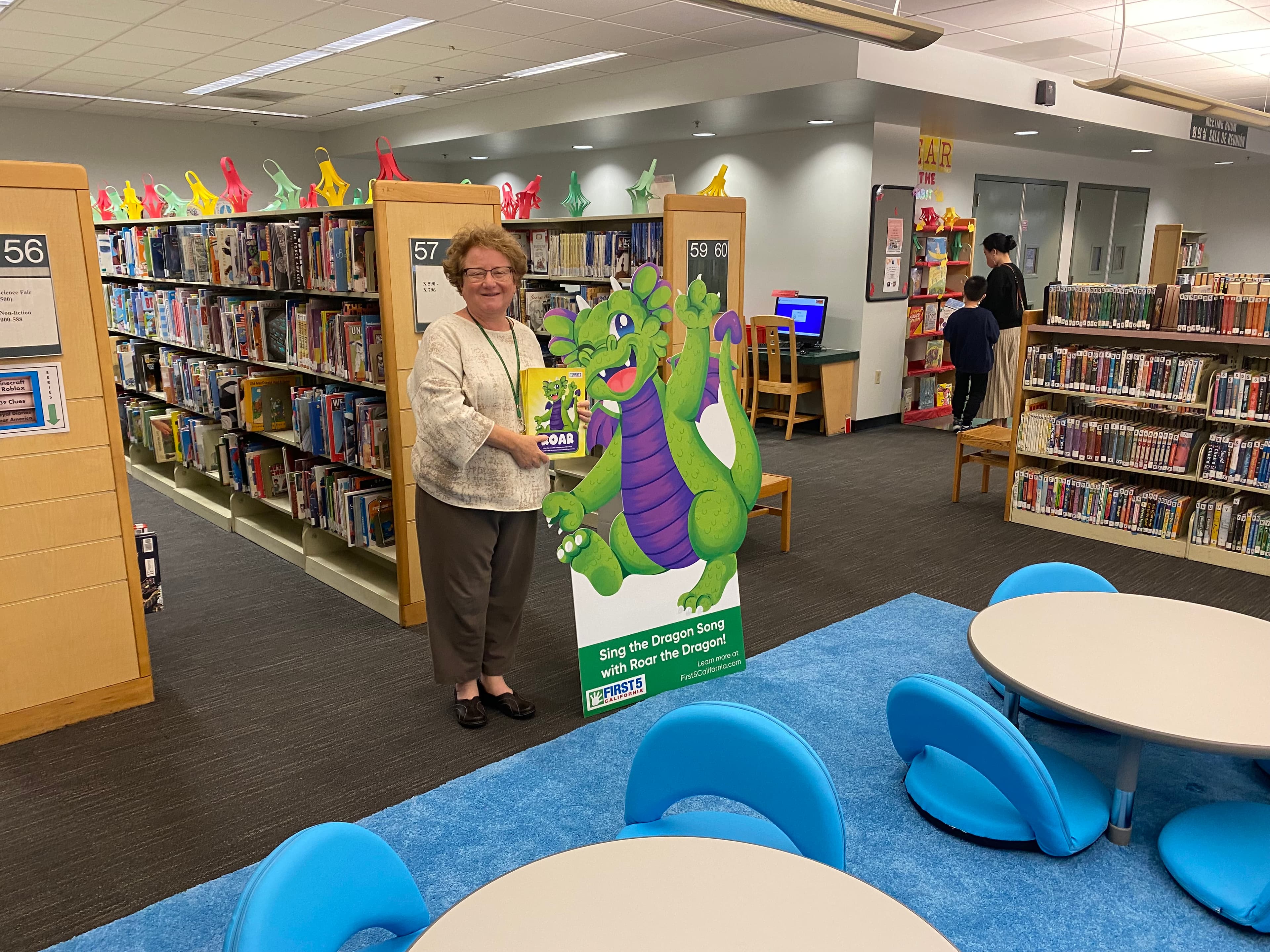 A librarian holding a children's book next to a custom dragon reading-program standee inside a public library