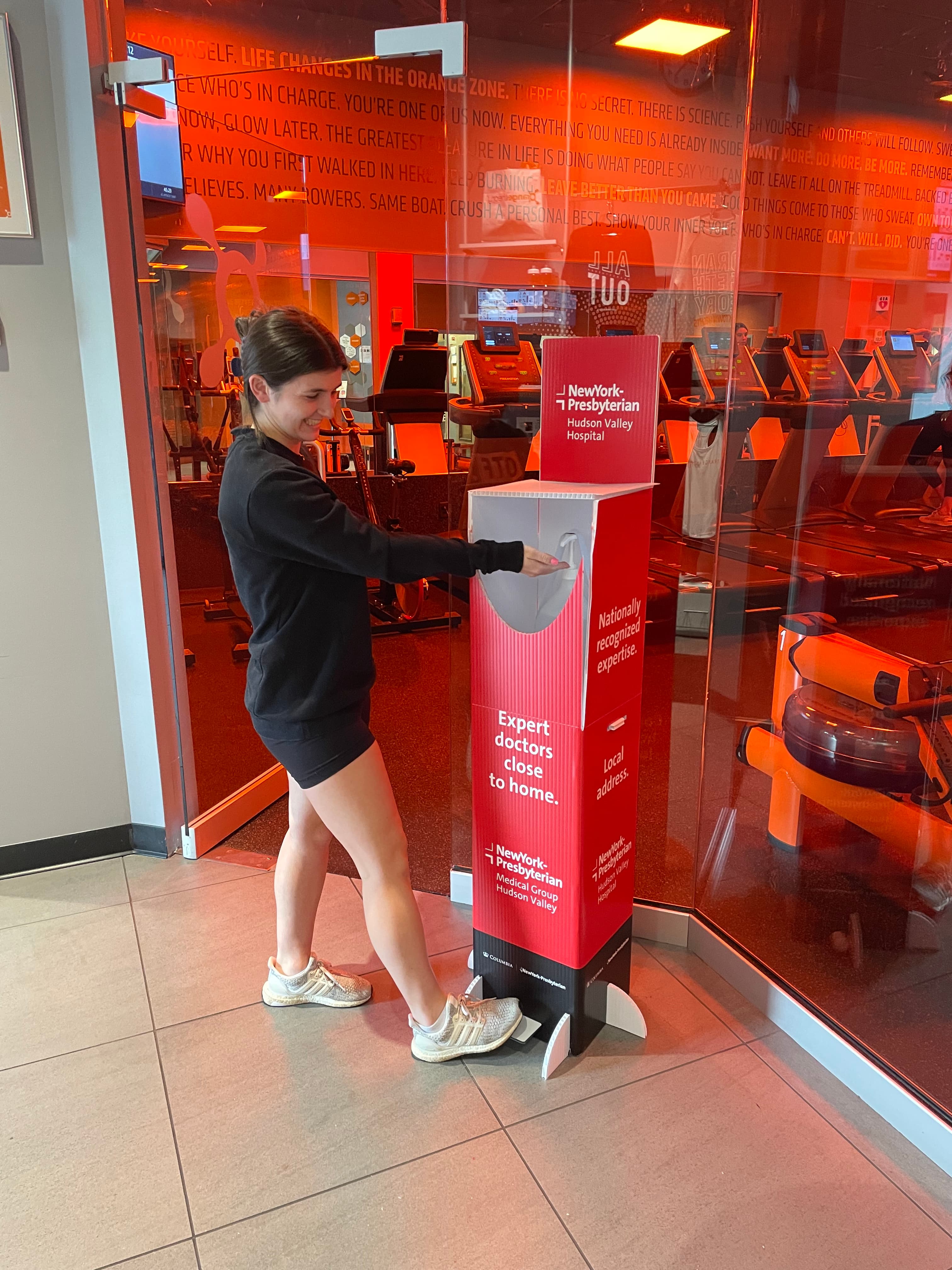 A gym member using a branded hand-sanitizer station beside an approved healthcare campaign display at an Orangetheory Fitness studio